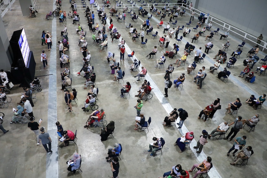 Dozens of people sit spaced out in a large hall awaiting vaccines