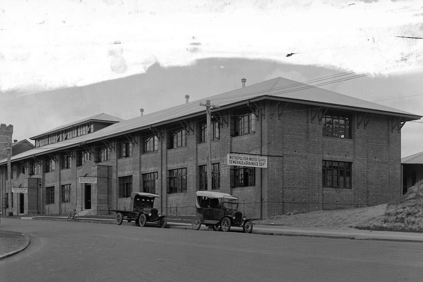 Black and white photo of two storey brick building with two vintage cars parked outside.