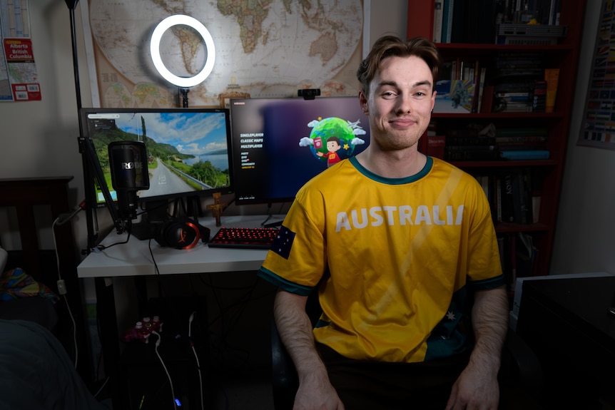 A man smiles with short brown hair wears a green and gold jersey, he sits in front of computer with map open
