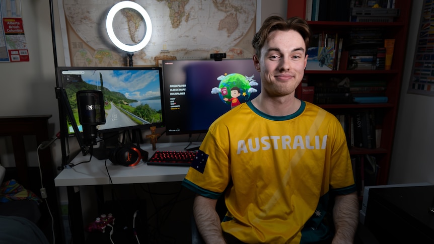 A man smiles with short brown hair wears a green and gold jersey, he sits in front of computer with map open