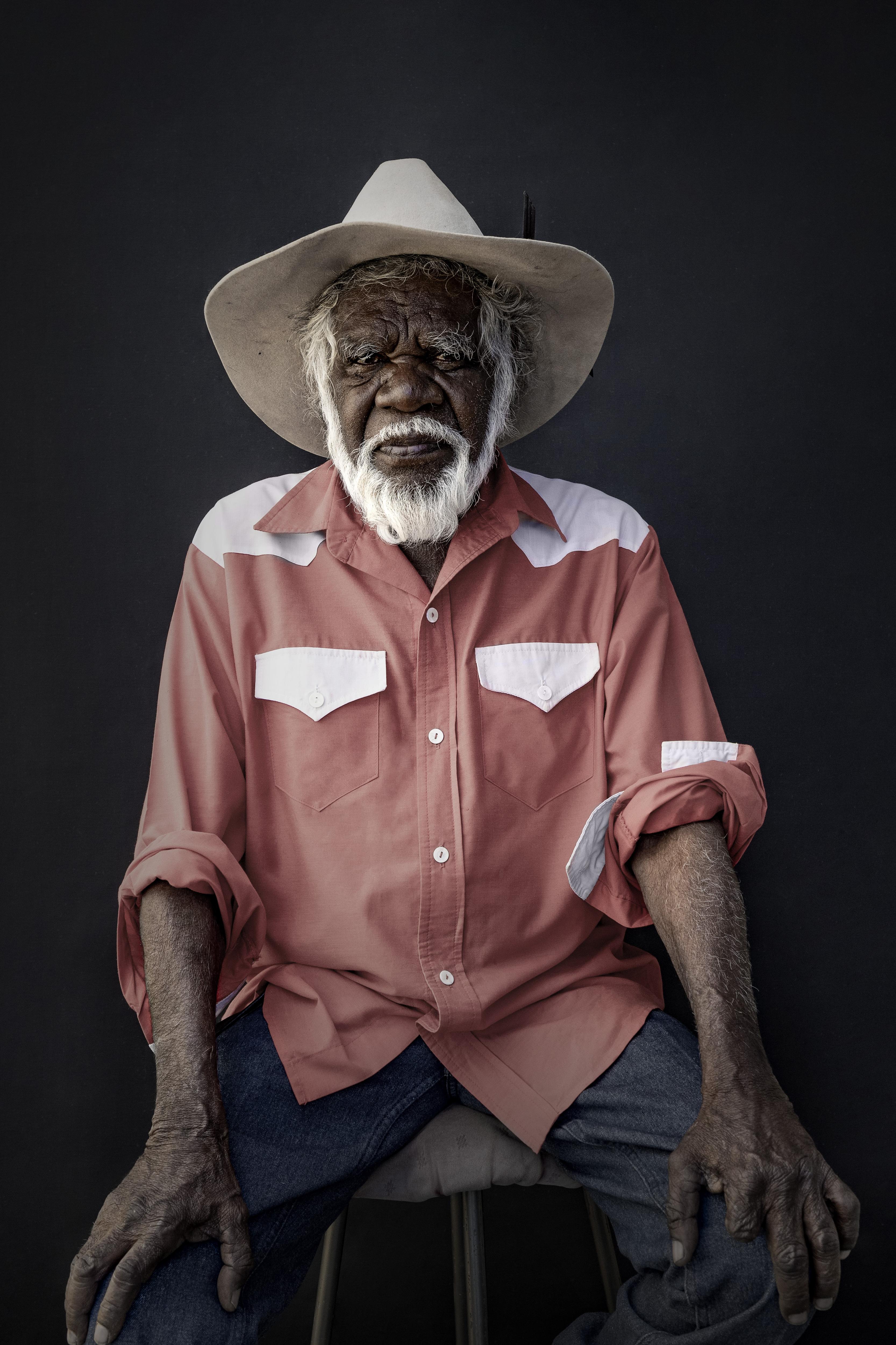 an indigenous man sits on a stool against a black backdrop earing orange button up shirt and white hat with a white beard