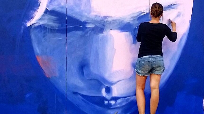 View from behind of an artist working on a large painting of a face on the outside wall of a building.