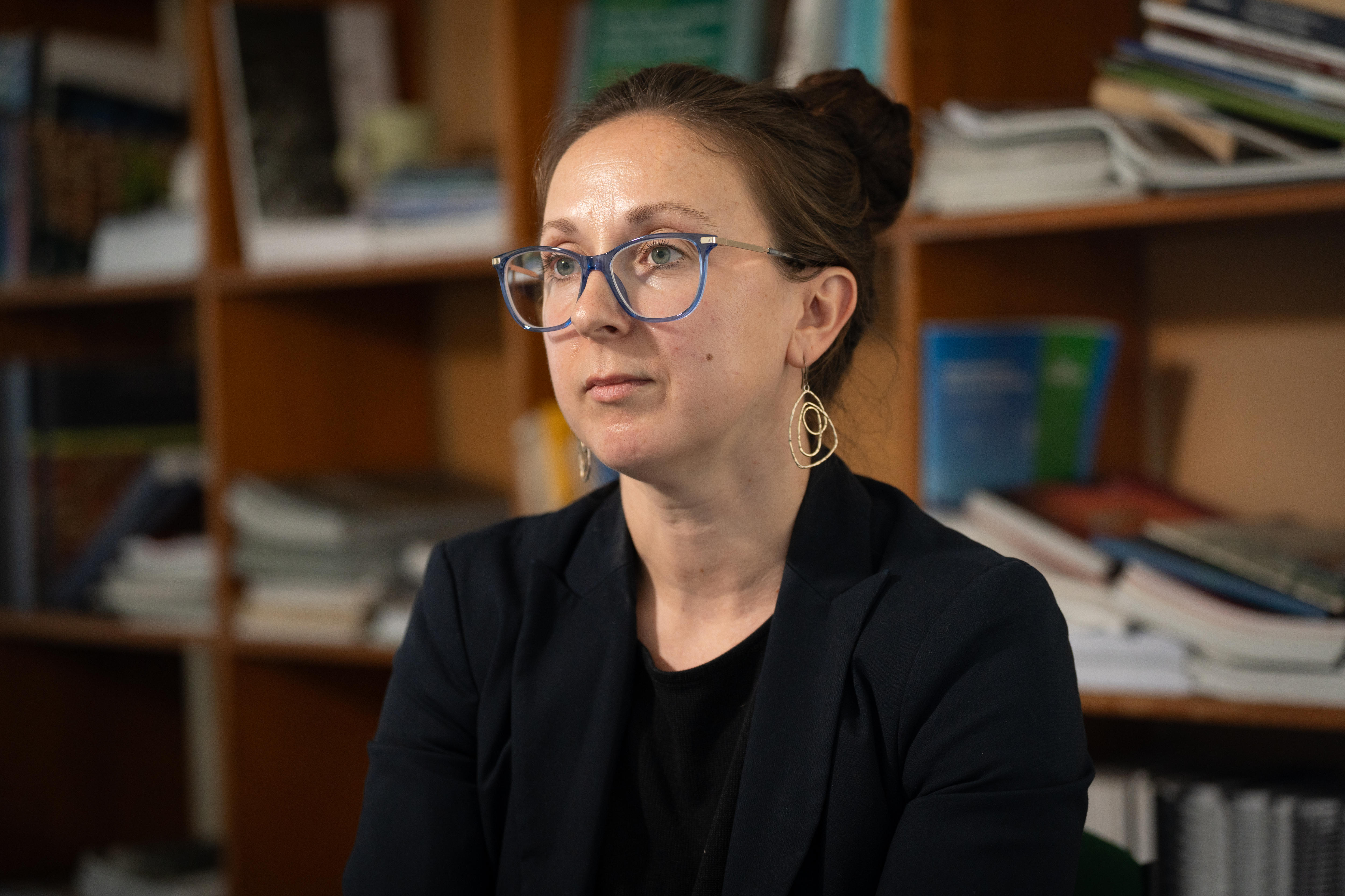 Una mujer blanca, con blusa y chaqueta negras, mira a la izquierda, a la cámara, con gafas de montura azul y una estantería borrosa detrás de ella.