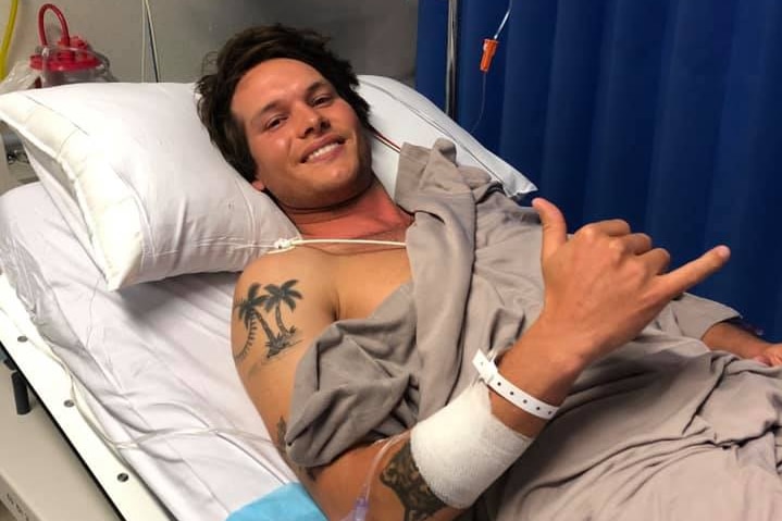 A man lies in a hospital bed making a 'hang loose' sign with one of his hands.