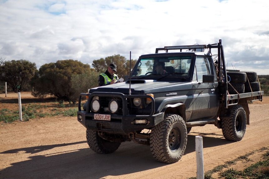 A police officer speaks to a man sitting in the front seat of a ute parked on a dirt road