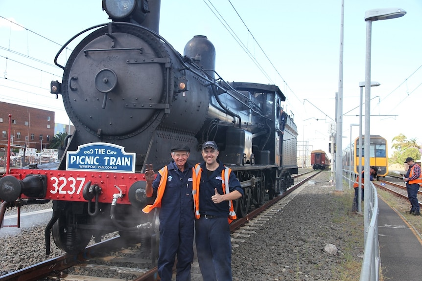 An older man and a younger man with thumbs up in front of a steam train