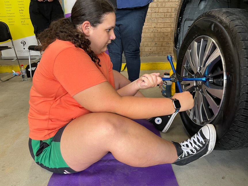 A girl in an orange t-shirt looks at the tyre and turns a four-pronged wrench.