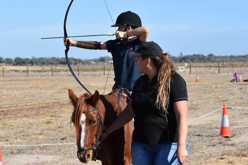 A young girl sits on a horse pulling an archery bow, guided by a woman standing up