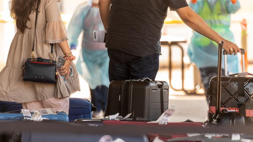 A man and a woman roll there luggage past people in PPE after arriving in Darwin from India.