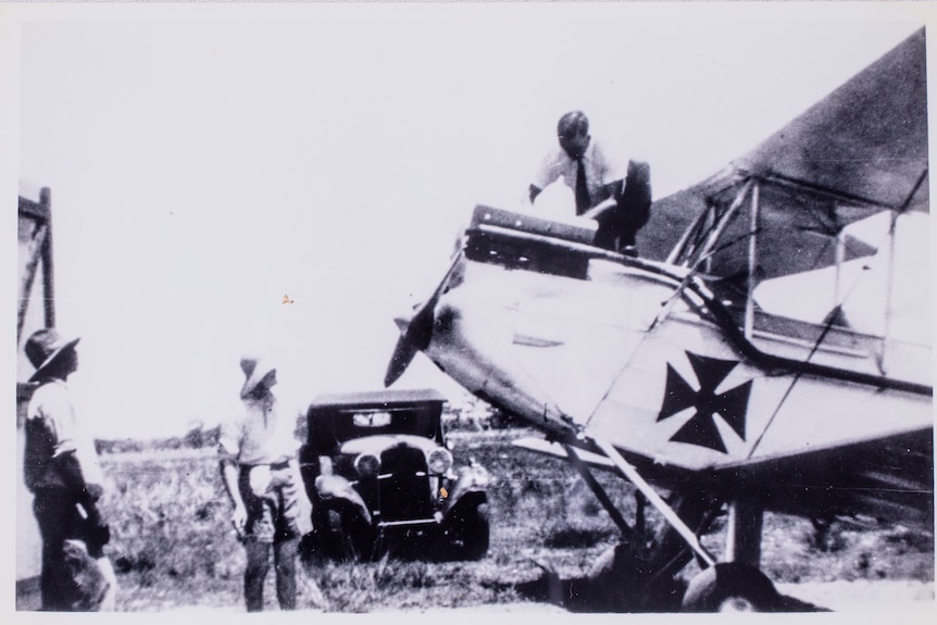 A man stands on top an old plane while two men look up at him. The photo is old and in black and white. 