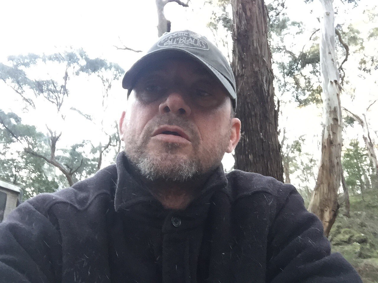 A selfie of a man in the bush wearing a cap and jumper with a beard. 