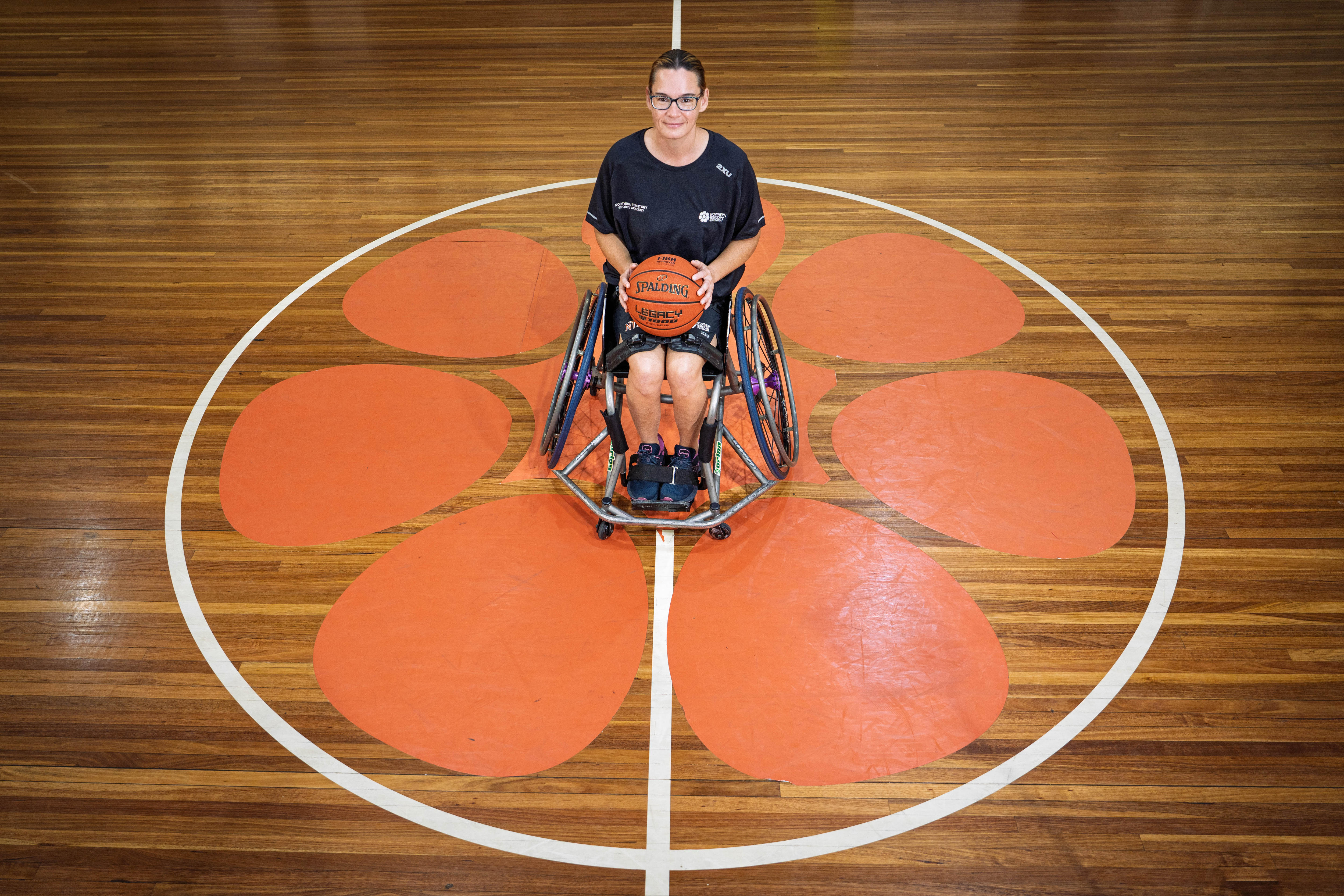 Vista de ángulo alto de una mujer sentada en silla de ruedas, baloncesto en mano, en el centro de la cancha de baloncesto con el logo de la flor frangipani