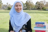 A teenager in a hijab sits on the grass by a books piled up to her shoulder. On her lap, she fingers through one book