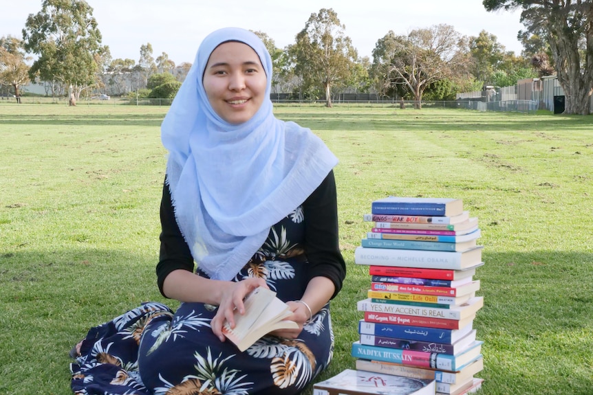 A teenager in a hijab sits on the grass by a books piled up to her shoulder. On her lap, she fingers through one book