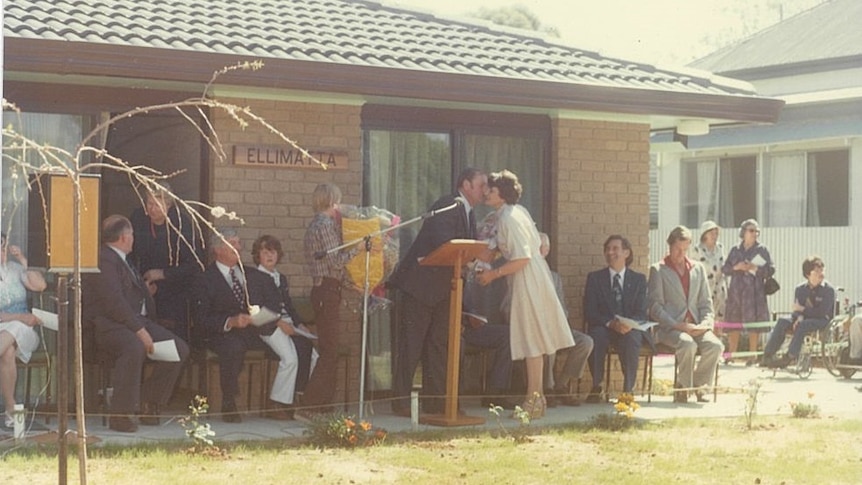 An old photo of a group of people stand and sit in front a brick house 