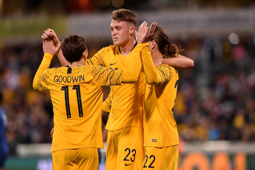 Harry Souter embraces and celebrates with Jackson Irvine and Craig Goodwin after a goal. They all wear yellow Socceroos jumpers