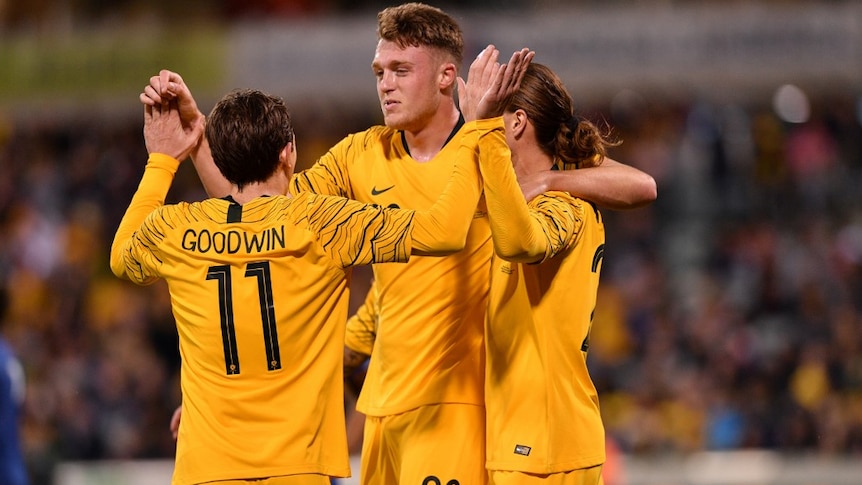 Harry Souter embraces and celebrates with Jackson Irvine and Craig Goodwin after a goal. They all wear yellow Socceroos jumpers