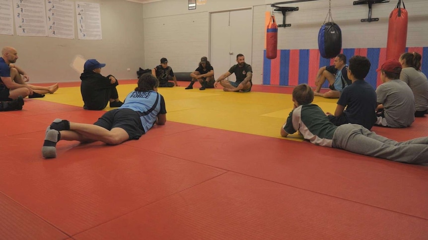 A group of about 12 youths sit in a circle on a yellow and red mat listening to a man.