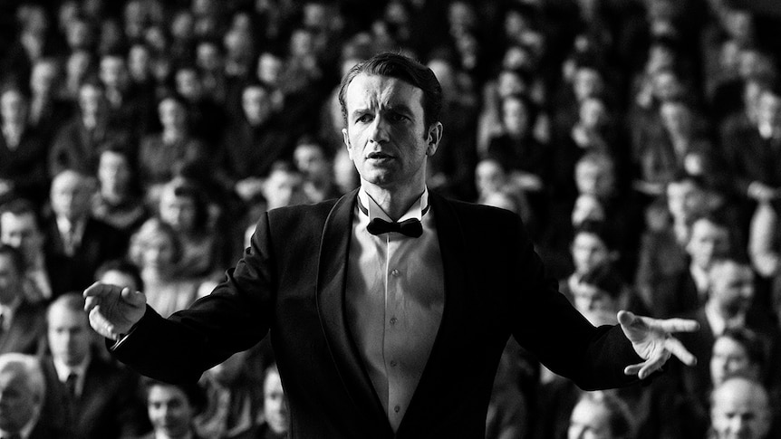 Black and white still of Tomasz Kot as conductor in front of audience in 2018 film Cold War.