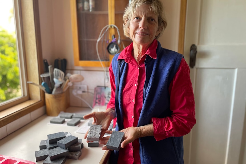 A woman stands holding two bars of charcoal soap with a pile of soap on the bench beside her.