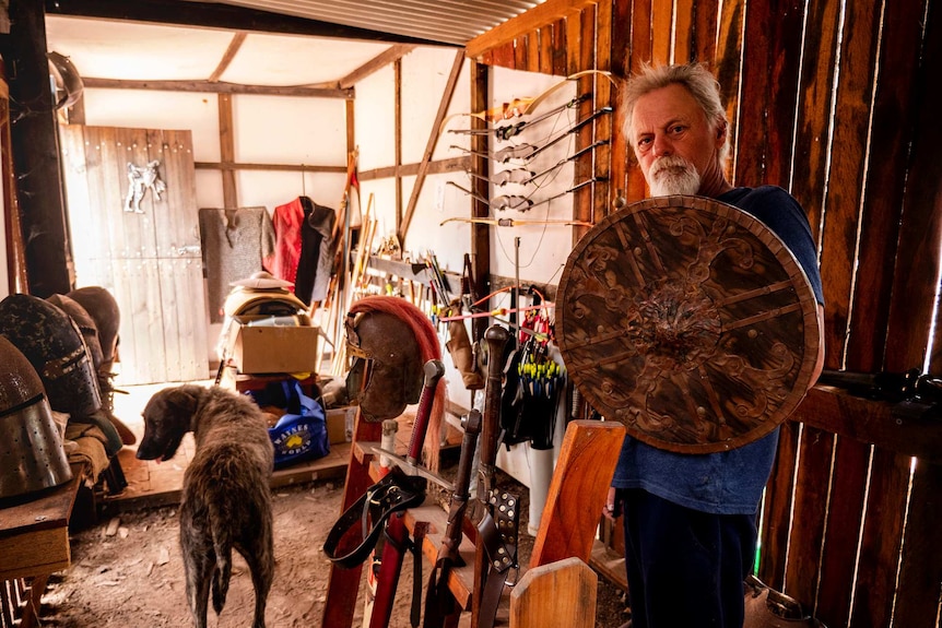 A man and large wolfhound dog stand in a timber room surrounded by bows, arrows, swords and helmets.