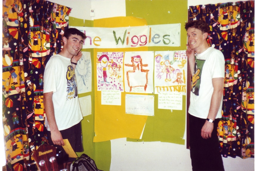 A 90s photo of two men in white tshirts standing next to a poster drawn by kids with the title The Wiggles