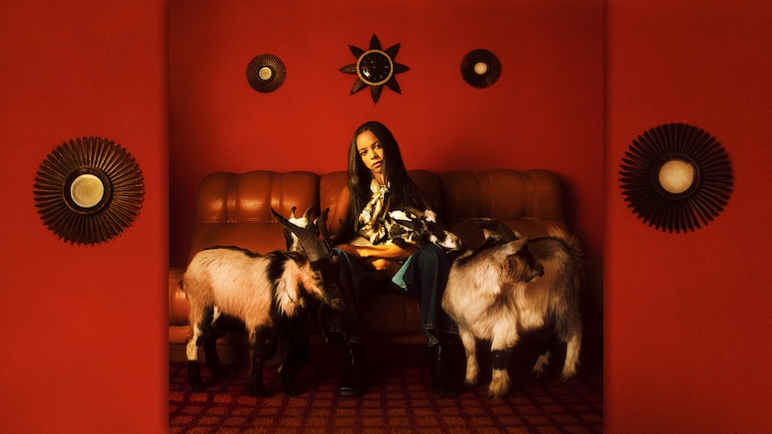 TSHA sits on a leather couch in a red room holding a goat on her lap. Two other goats stand on either side of her.