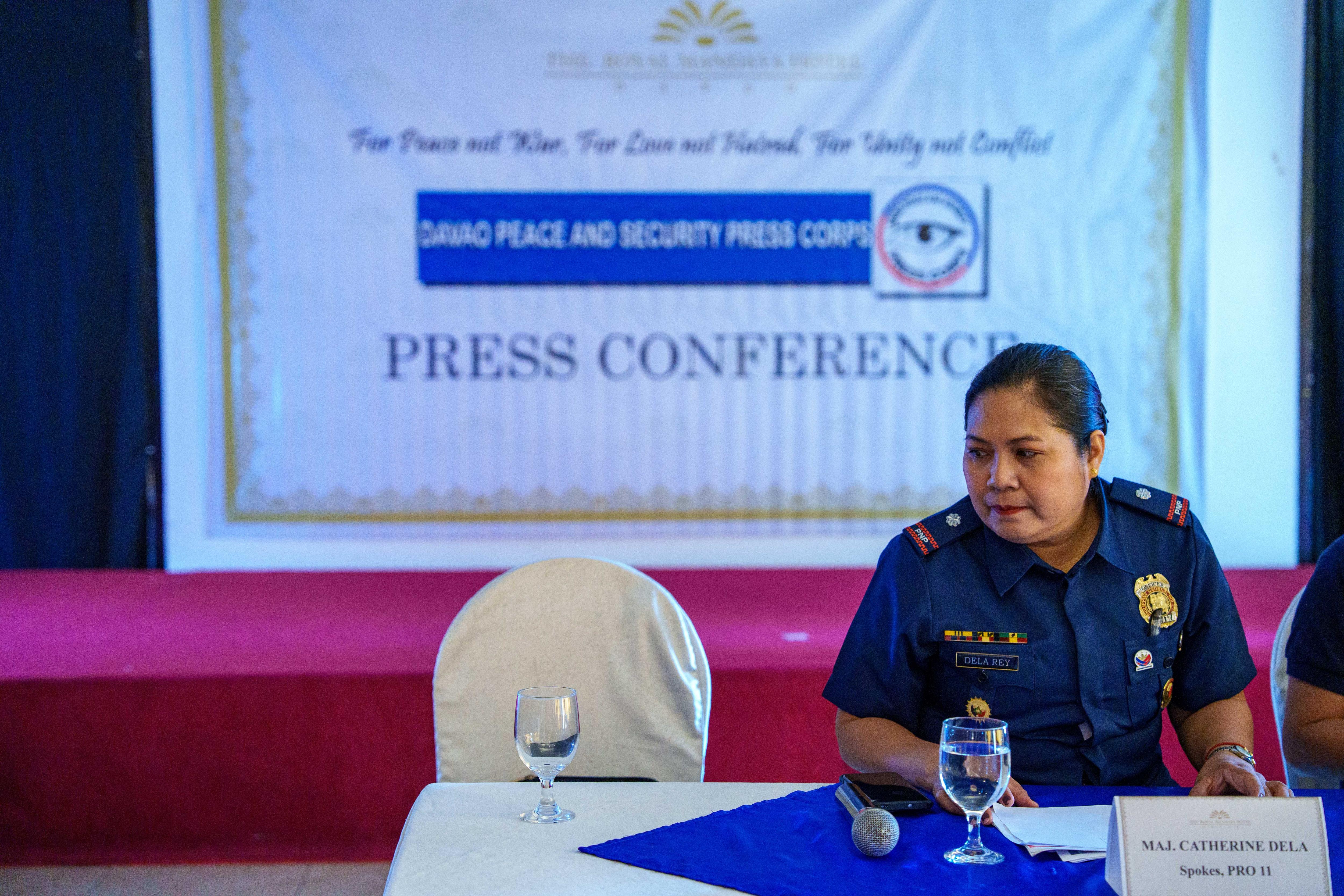 Uma policial sentada a uma mesa, em frente a uma placa que diz CONFERÊNCIA DE IMPRENSA.