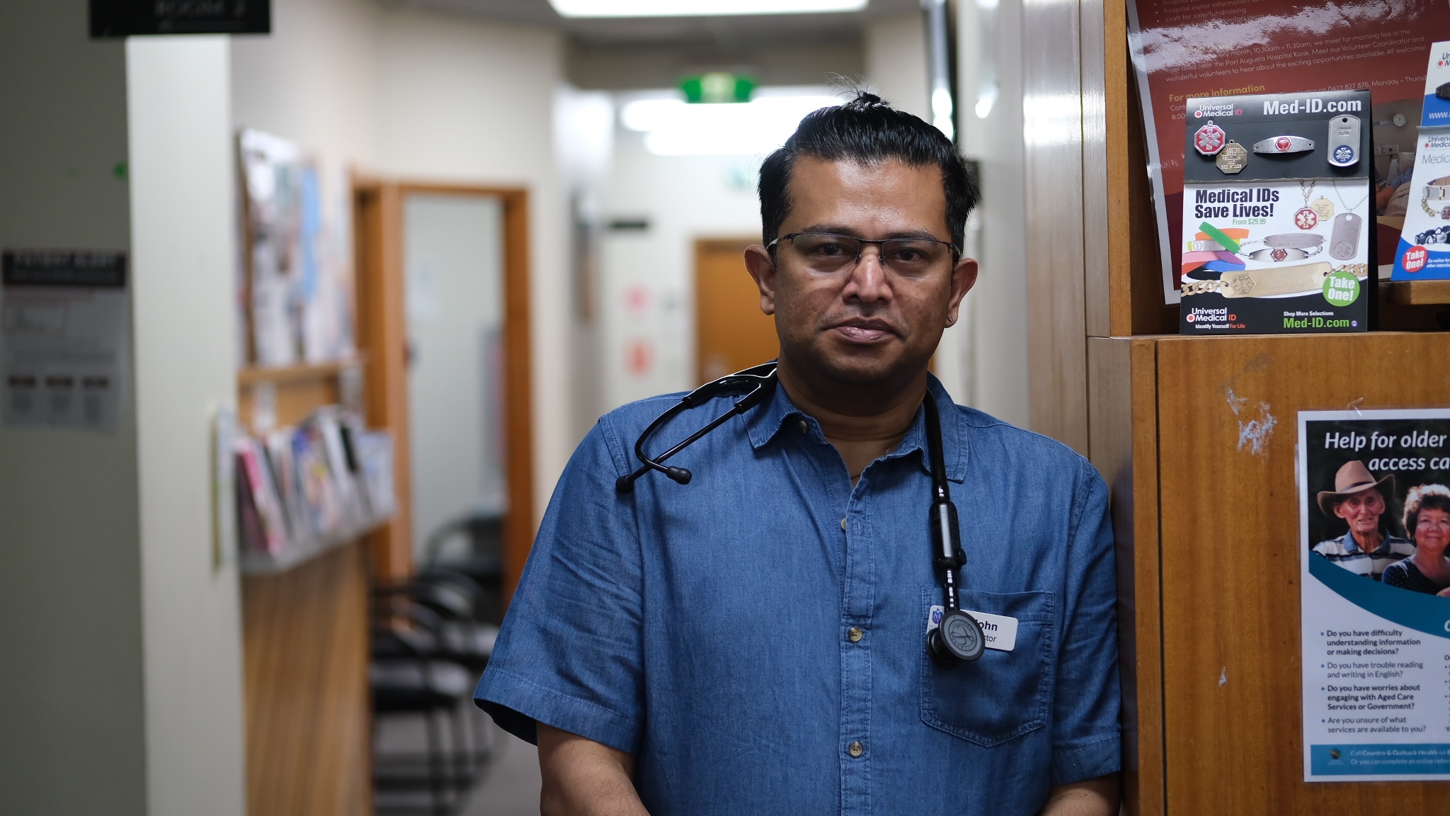 Um homem com um estetoscópio pendurado no pescoço fica em frente a armários dentro de um centro médico.
