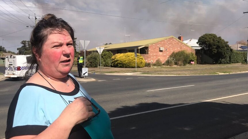 A woman stands in front of a residential street as smoke rises behind a house in the background.