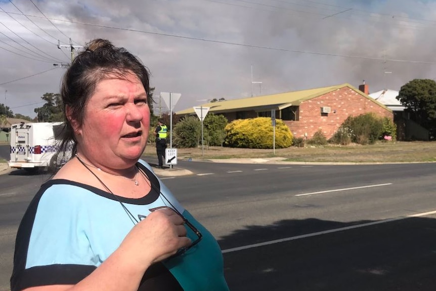 A woman stands in front of a residential street as smoke rises behind a house in the background.
