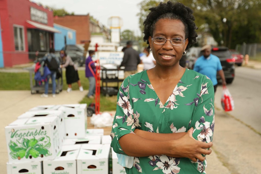 A black woman wearing silver glasses and a green shirt with flowers stands smiling with her arms crossed.