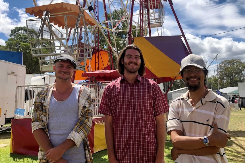 Three young men standing in front of a Ferris wheel smiling.