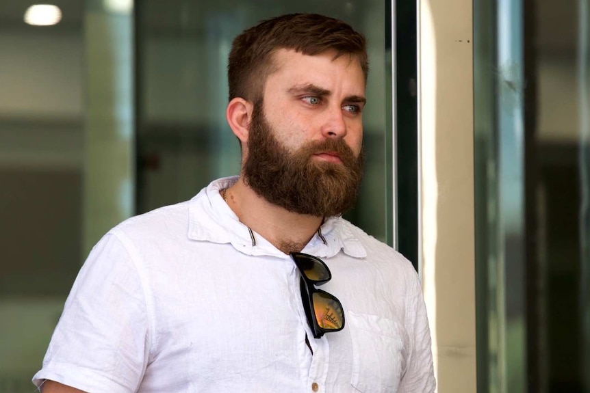 A mid-shot of a young man with a beard and a white shirt exiting a building.