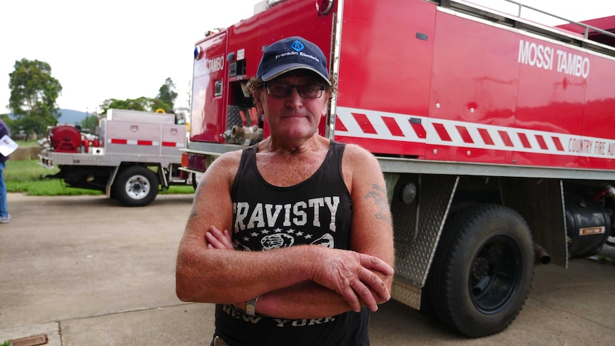 An older man wearing a cap, glasses and singlet with his arms crossed looking at the camera.