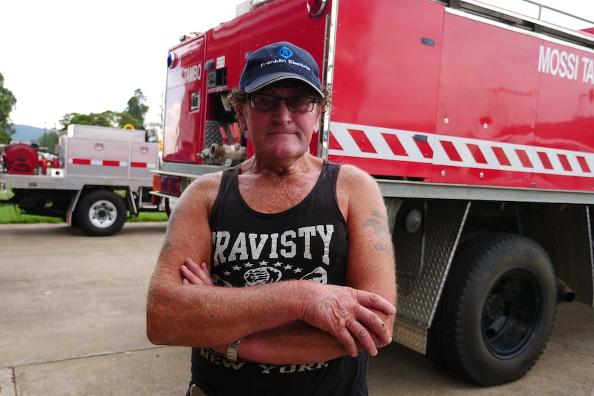 An older man wearing a cap, glasses and singlet with his arms crossed looking at the camera.