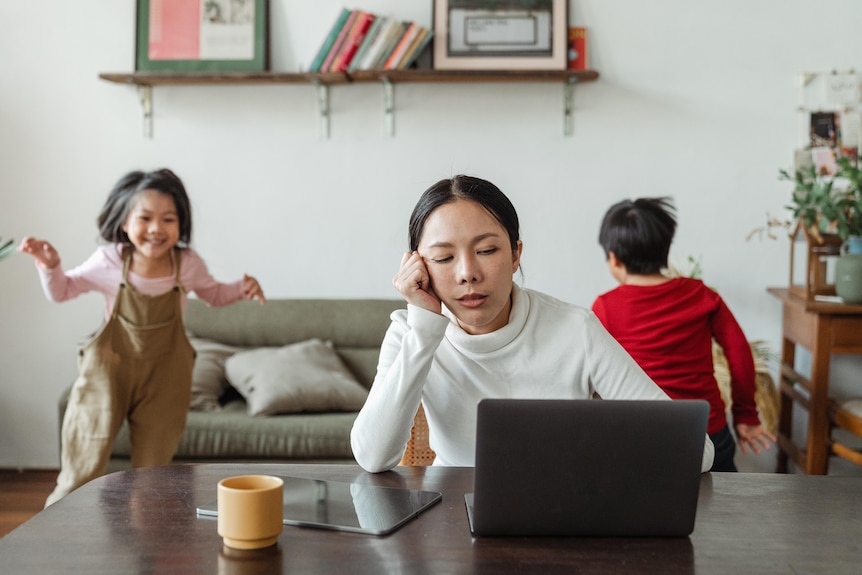 A woman sits in front of her computer with her head in her hand while two children play behind her. 