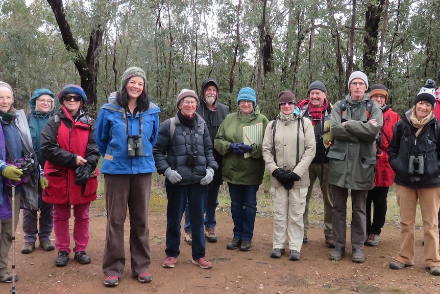 A group of people smile, standing outside wearing jackets and beanies, holding binoculars.