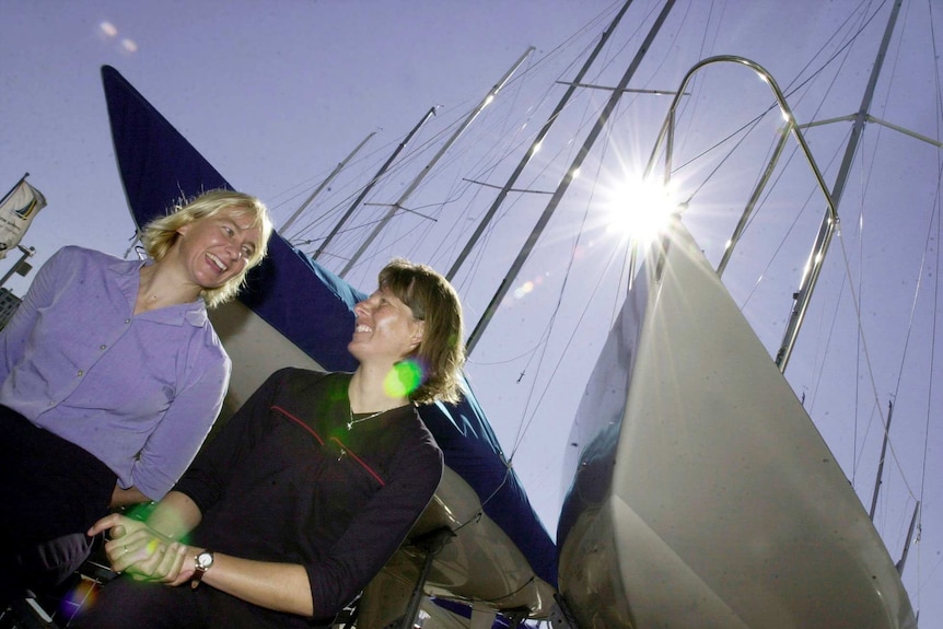 Two women look at each other and smile in front of sailing boats at a marina.