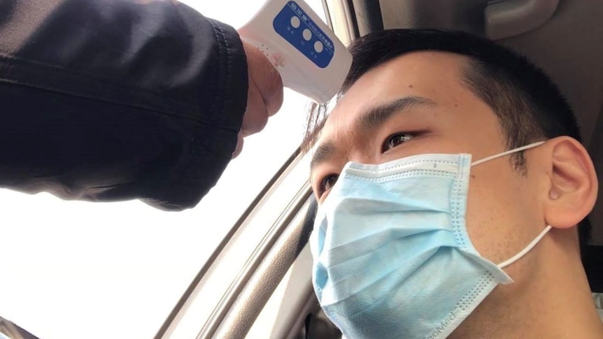 A man in a mask in a car is checked with a thermometer, being held through his car window.