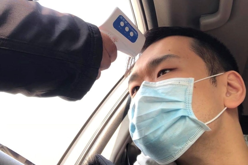 A man in a mask in a car is checked with a thermometer, being held through his car window.