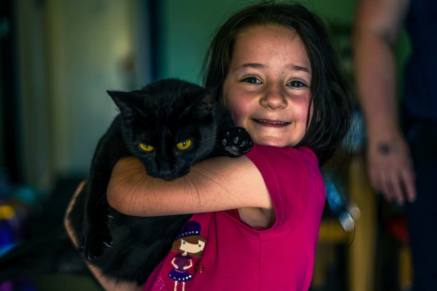 A young girl smiles for a photo with a black cat clutched in her arms.