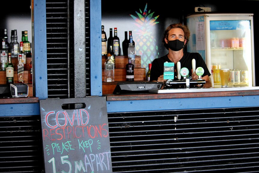 Un hombre con una máscara se para detrás de un bar en un albergue para mochileros junto a un letrero que le dice a la gente que se mantenga a distancia social