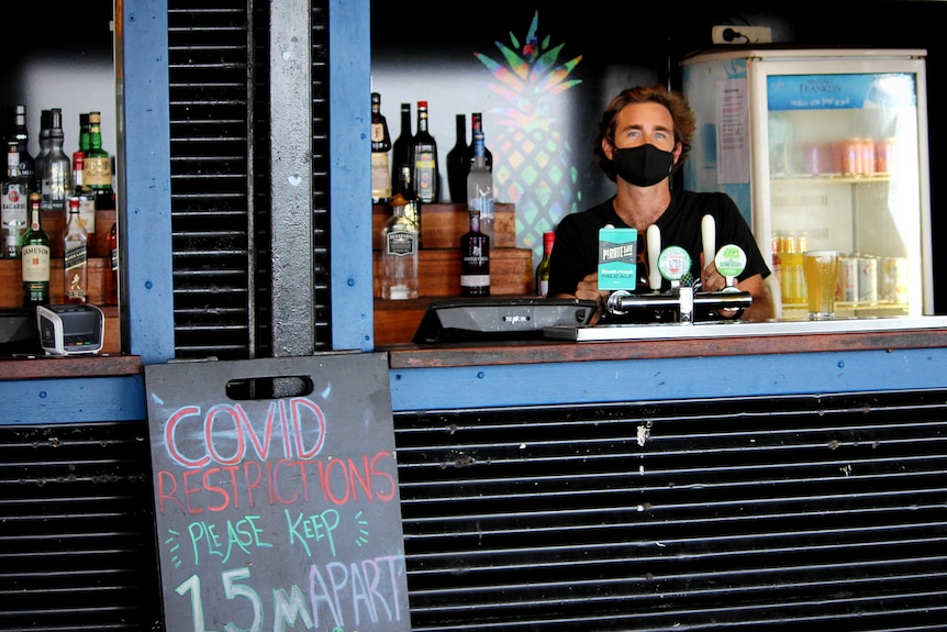 A man wearing a mask stands behind a bar at a backpackers hostel next to a sign telling people to social distance