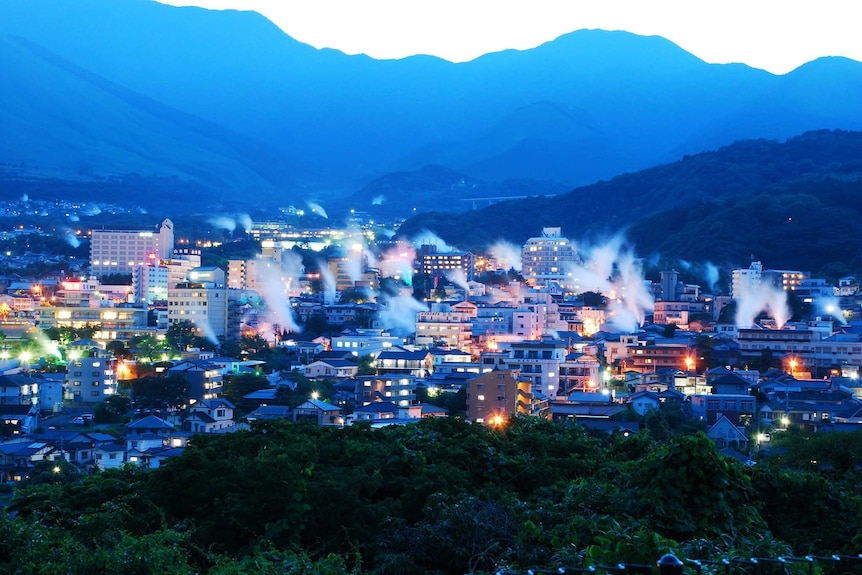 A wide sweeping view of buildings with white steam rising from various locations. This is set against a mountainous backdrop.