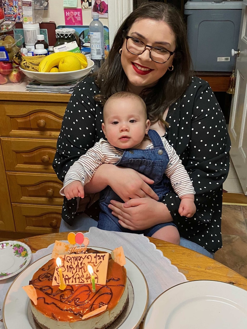 Natasha Biggins holds her baby as she sits at a table with a birthday cake on it.