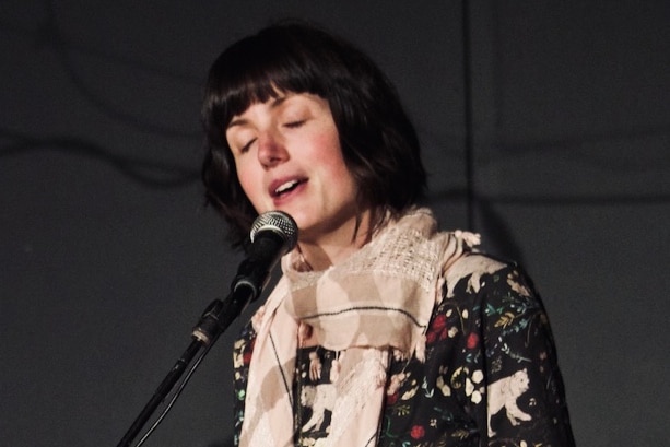 A woman with dark hair stands singing at a microphone with her eyes closed.