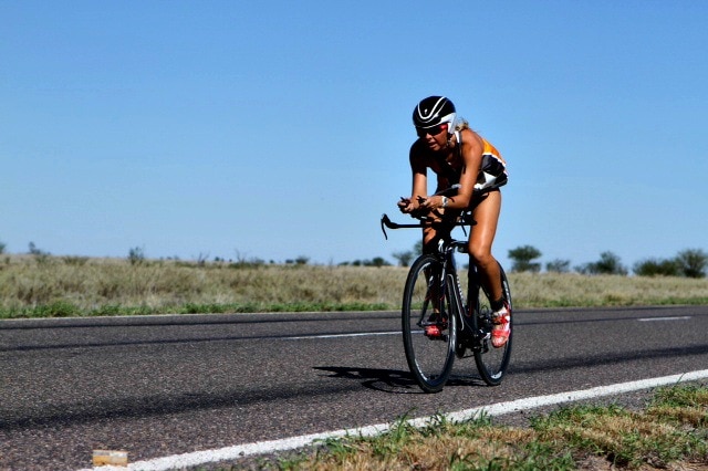 A cyclist rides along a bitumen road with dry grass on either side of the road and the sun bearing down from above.