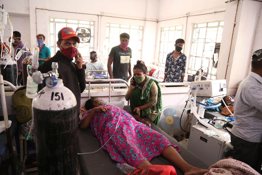 Patients laying on beds use oxygen cylinders in hospital as relatives stand over them.