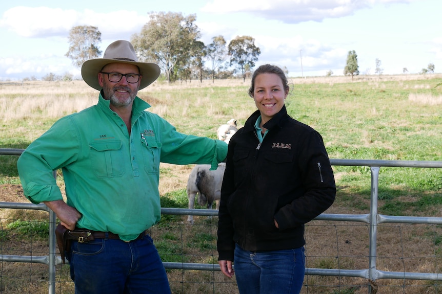 Man with a hat on and green work shirt stands smiling next to a women with a black jacket on. Both stand infront of a gate.